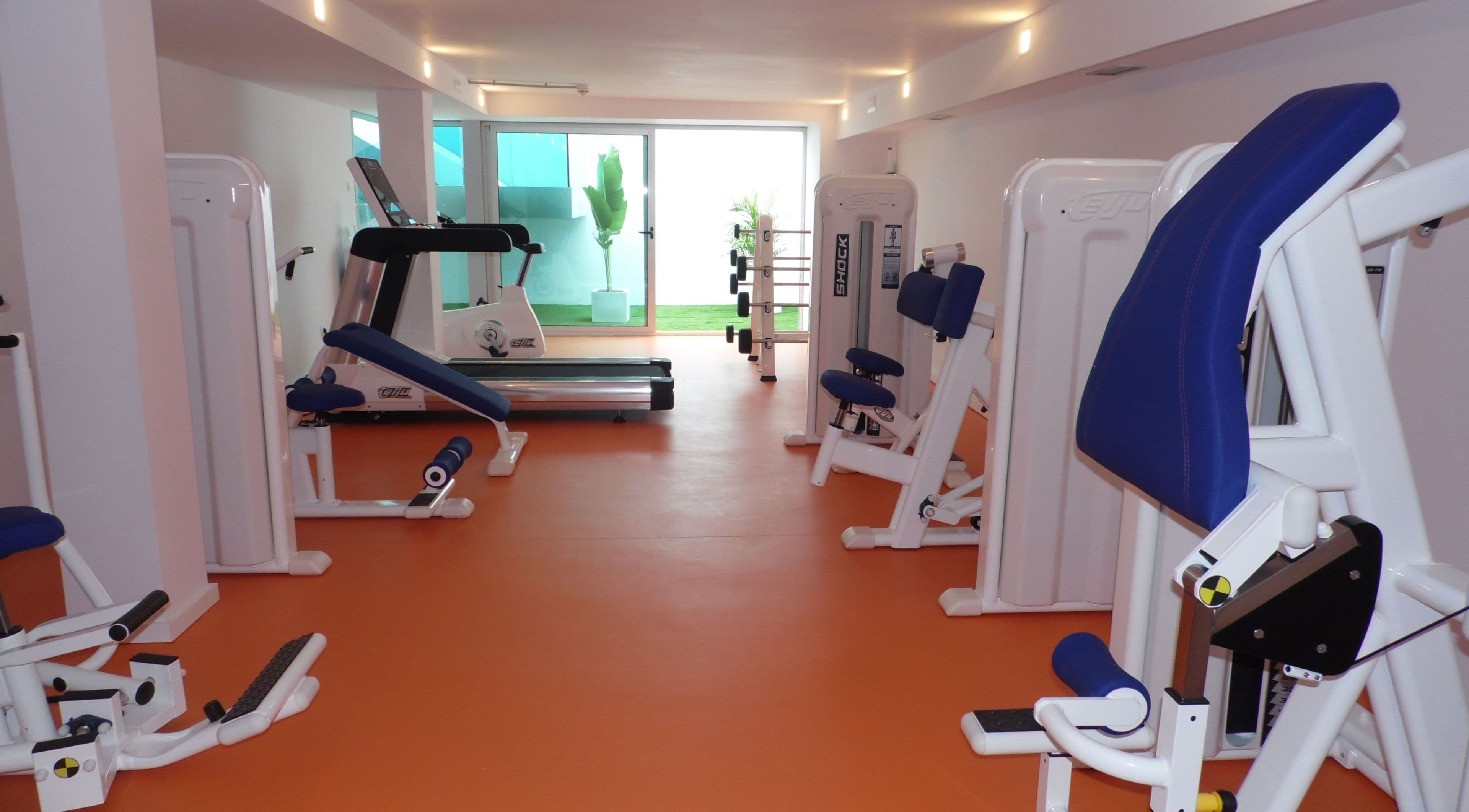 A gym with various exercise equipment, including treadmills and weight machines, arranged in a spacious room with orange flooring.