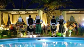 Zona de piscina con un grupo de personas vestidas con atuendo tradicional, una palmera y un edificio con área de estar cubierta.