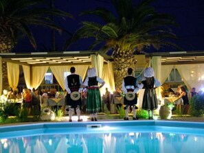 Un espace aménagé au bord de la piscine, où se trouve un groupe de personnes vêtues de tenues traditionnelles, un palmier et un bâtiment doté d’un espace de repos abrité.