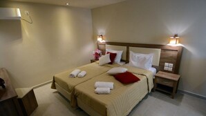 Una cama doble con una colcha color beige, dos toallas blancas y una almohada roja. Hay dos mesitas de noche con lámparas, un aparador de madera y un equipo de aire acondicionado instalado en la pared.