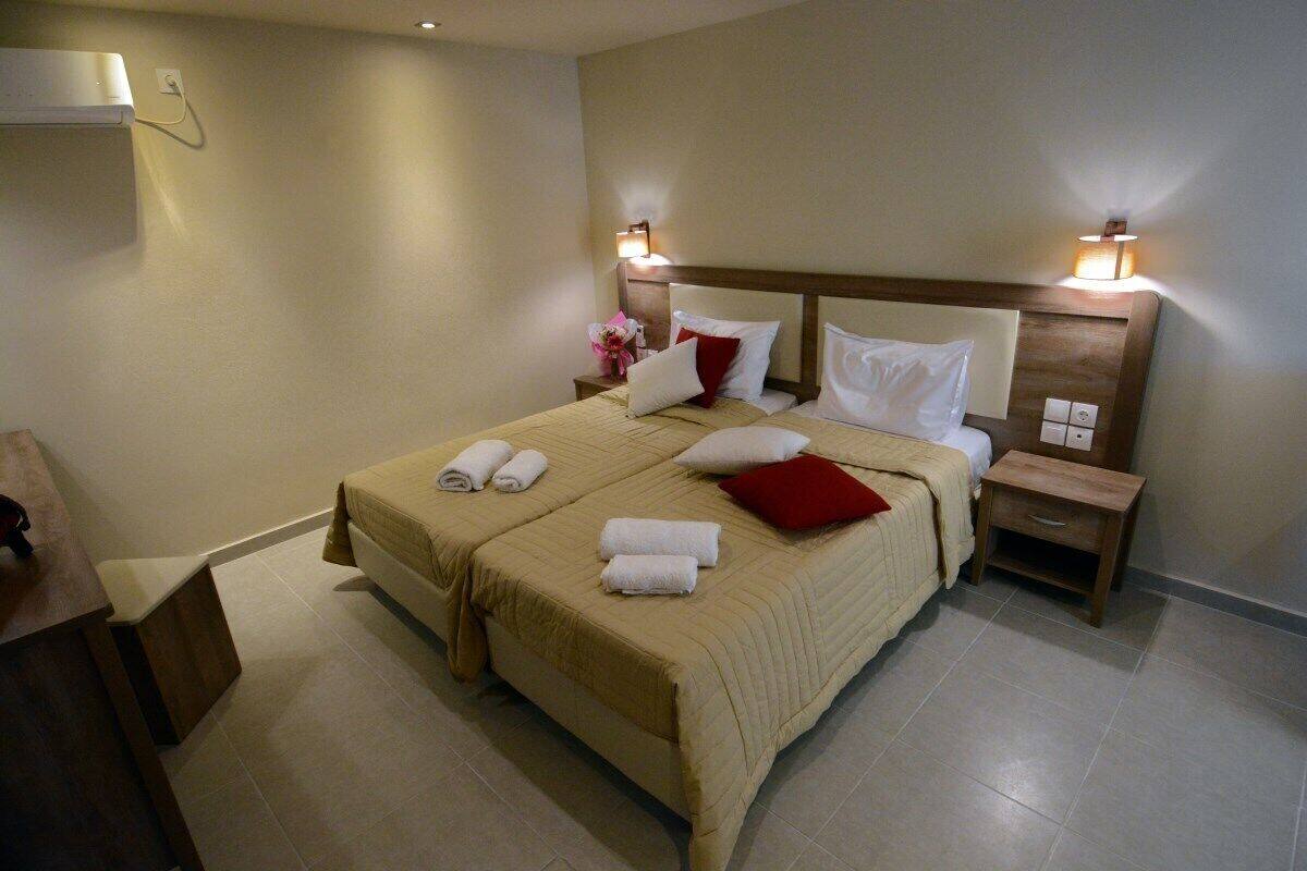 Una cama doble con una colcha color beige, dos toallas blancas y una almohada roja. Hay dos mesitas de noche con lámparas, un aparador de madera y un equipo de aire acondicionado instalado en la pared.