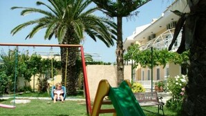 Un parque infantil con tobogán y columpios, rodeado de palmeras y un edificio al fondo.