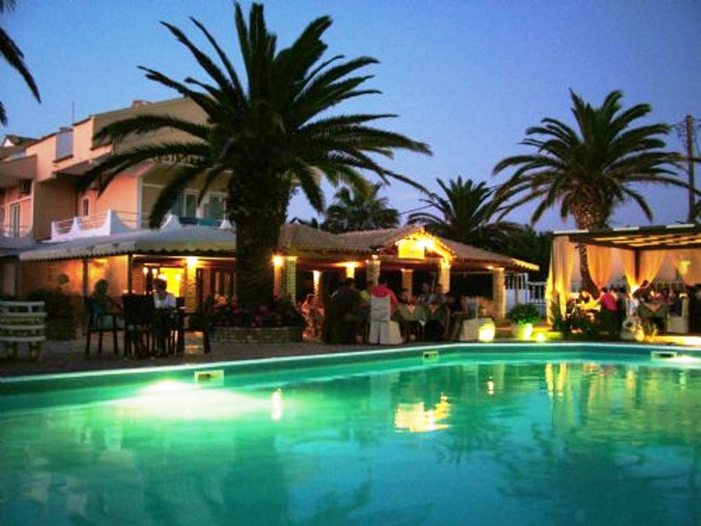 Zona de piscina con palmeras, un edificio con asientos al aire libre y un bar junto a la piscina.