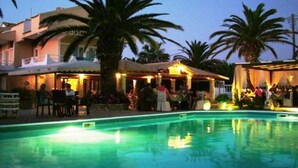 Zona de piscina con palmeras, un edificio con asientos al aire libre y un bar junto a la piscina.