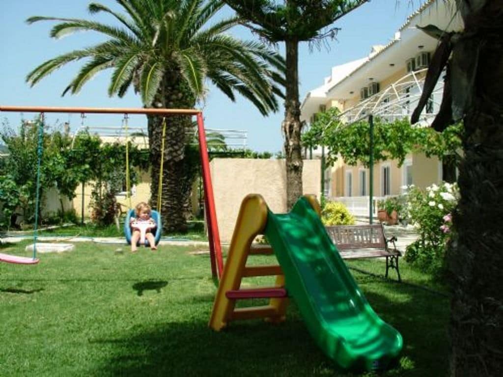 Un parque infantil con tobogán y columpios, rodeado de palmeras y un edificio al fondo.