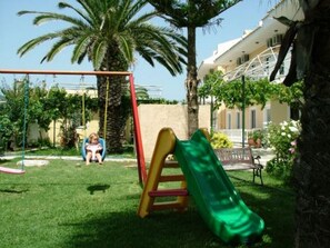 Ein Spielplatz mit Rutsche und Schaukeln, umgeben von Palmen und einem Gebäude im Hintergrund.