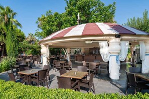 An outdoor café with tables and chairs, surrounded by greenery and a large umbrella structure. - Palmeras Beach Hotel (Alanya)