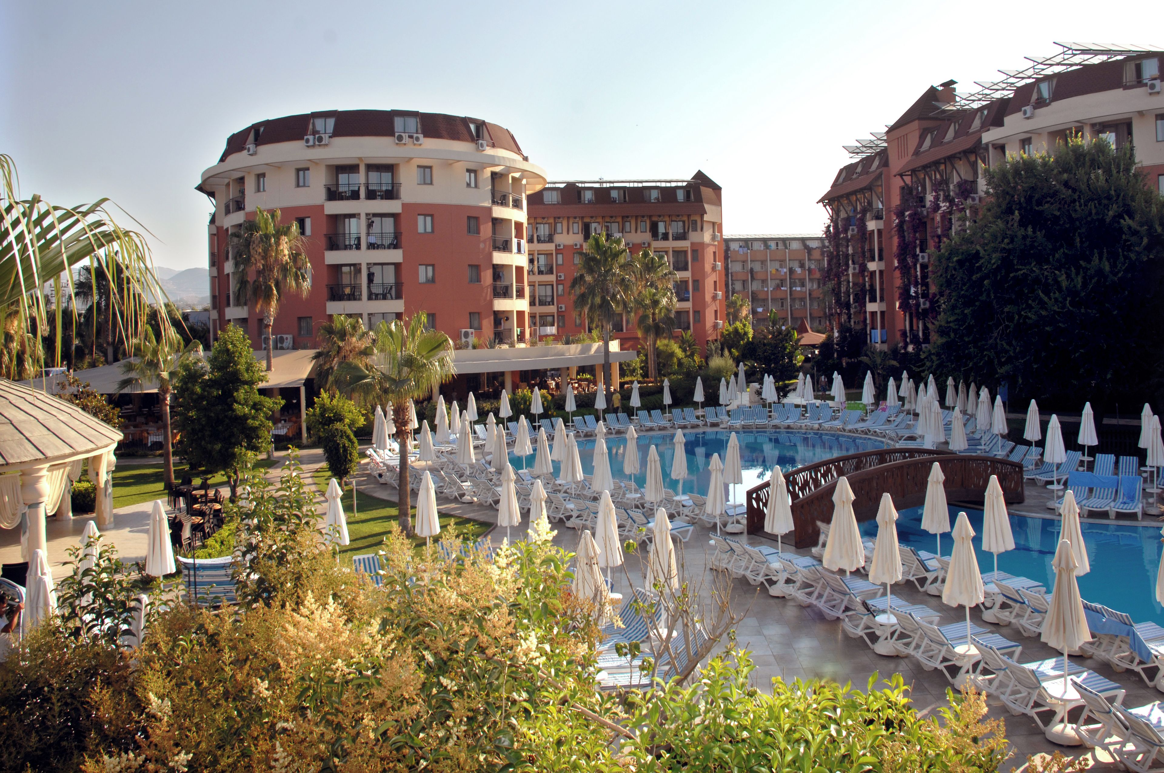 Ein Resort mit Swimmingpool, umgeben von Liegestühlen und Sonnenschirmen, mit mehrstöckigen Gebäuden im Hintergrund.