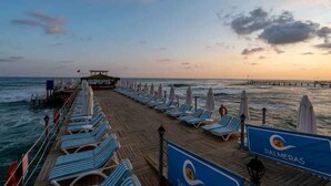 A wooden pier with lounge chairs and umbrellas, leading to a beach with a view of the ocean and a pier in the distance. - Palmeras Beach Hotel (Alanya)