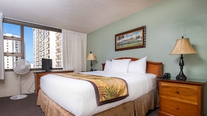 A hotel room with a large bed, two bedside tables, a nightstand, a desk with a computer, a standing fan, and a window with curtains.