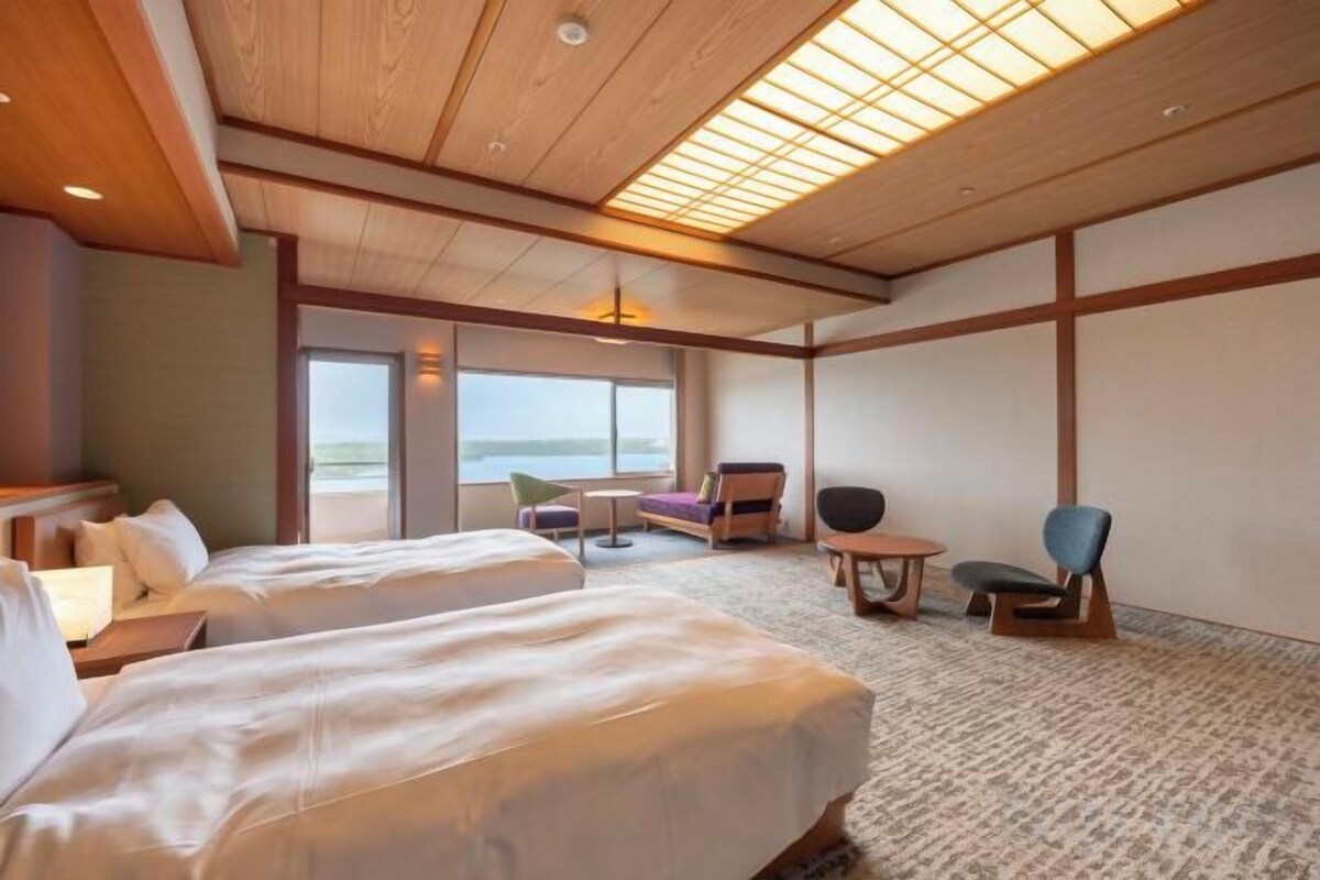 A hotel room with two beds, a seating area with a table, and large windows offering a view.