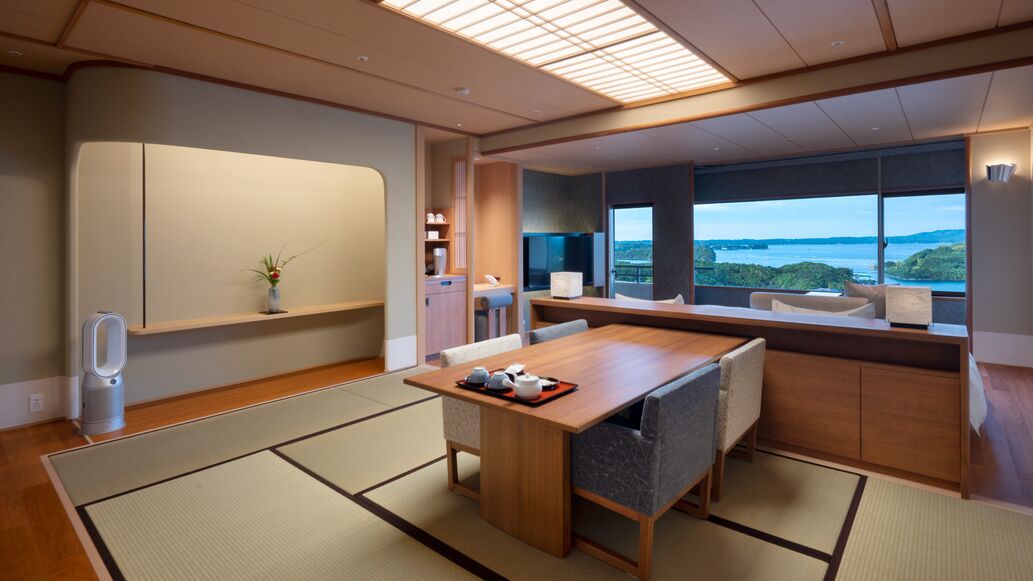 A modern hotel room with tatami flooring, a wooden dining table, a large window with a view, and a traditional Japanese aesthetic.