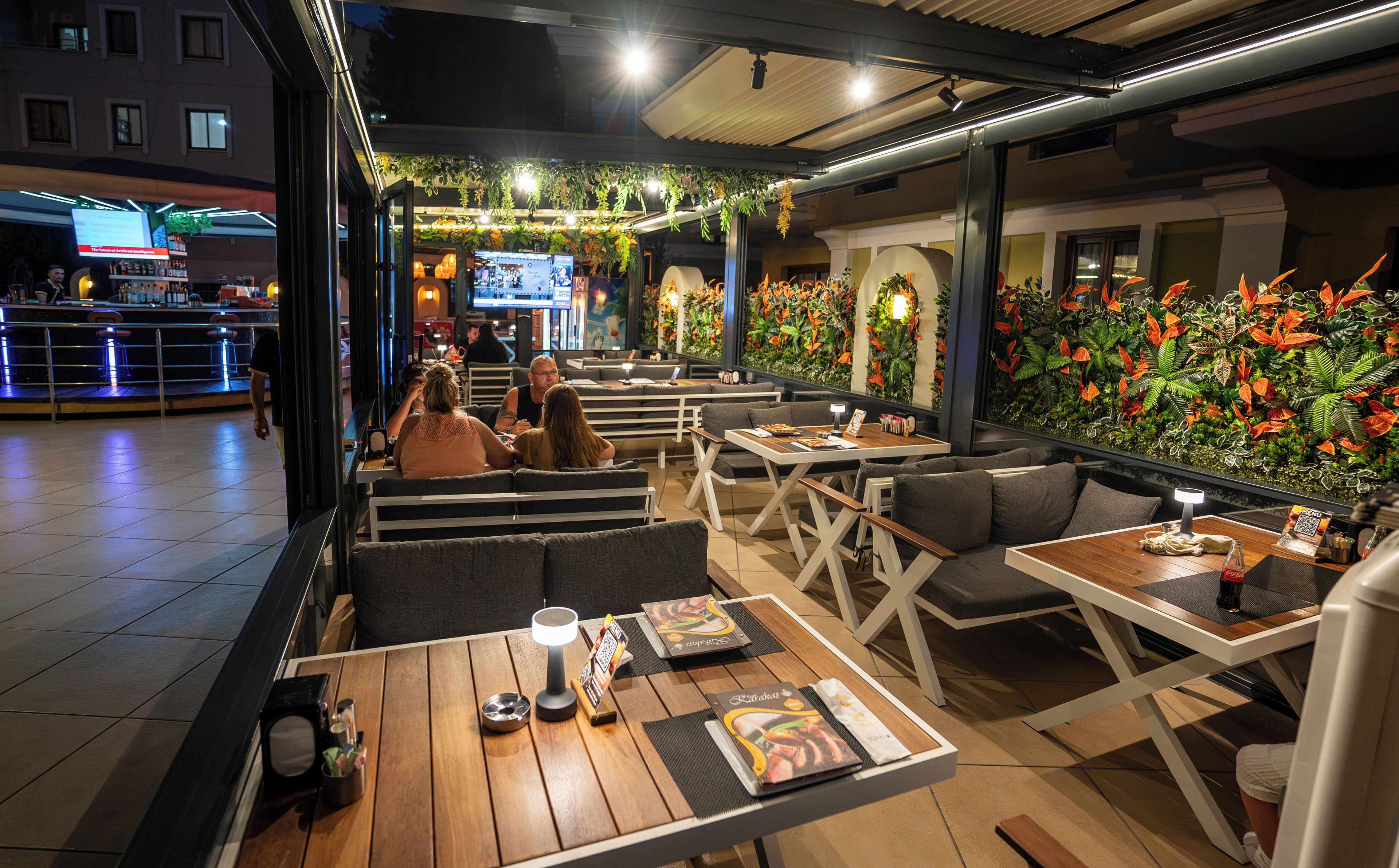 Un espacio de comedor al aire libre con mesas y bancos, un bar al fondo y una pared decorada con follaje artificial.