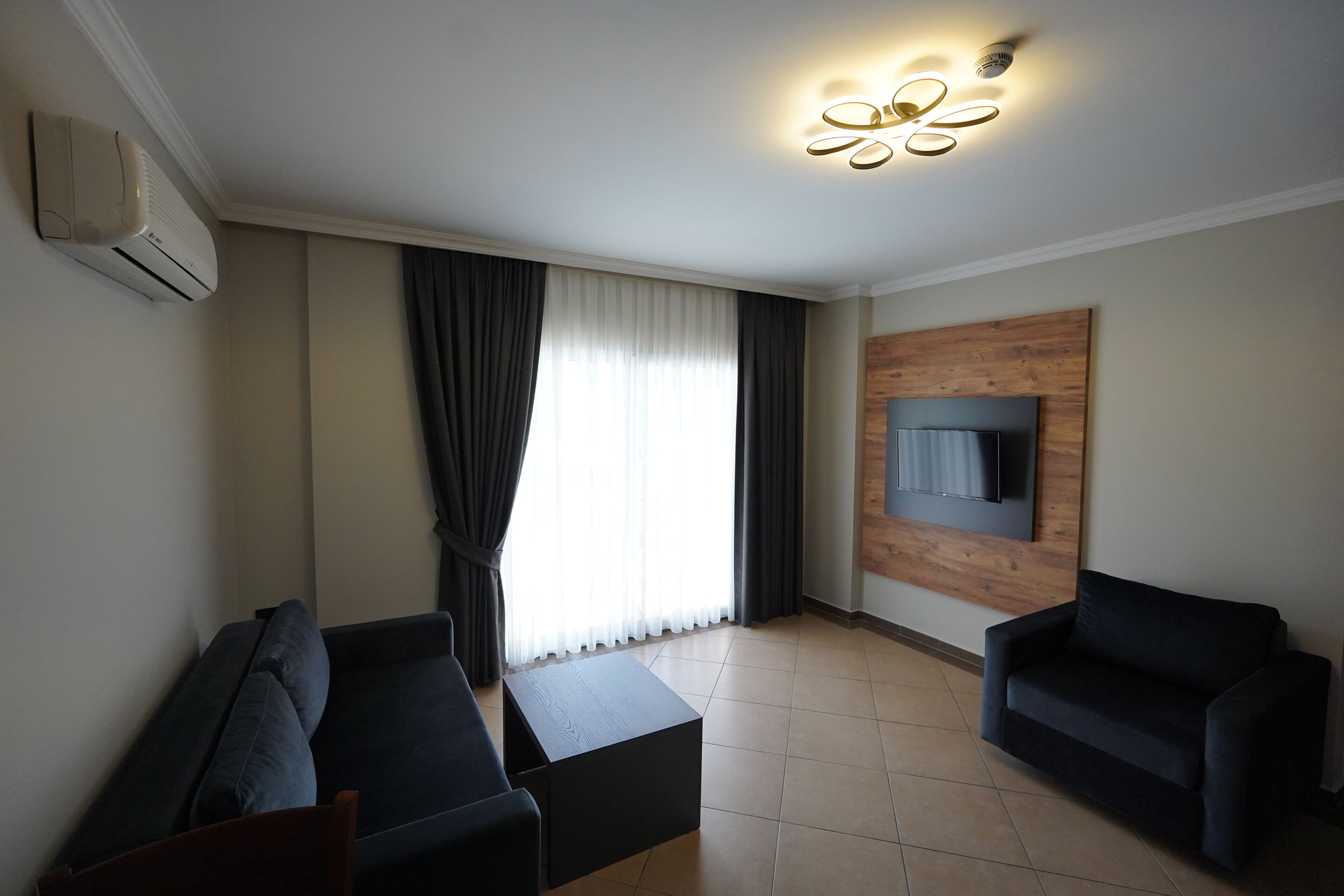 A modern living room with a sofa, a coffee table, a flat-screen TV mounted on a wooden panel, and a ceiling light fixture.