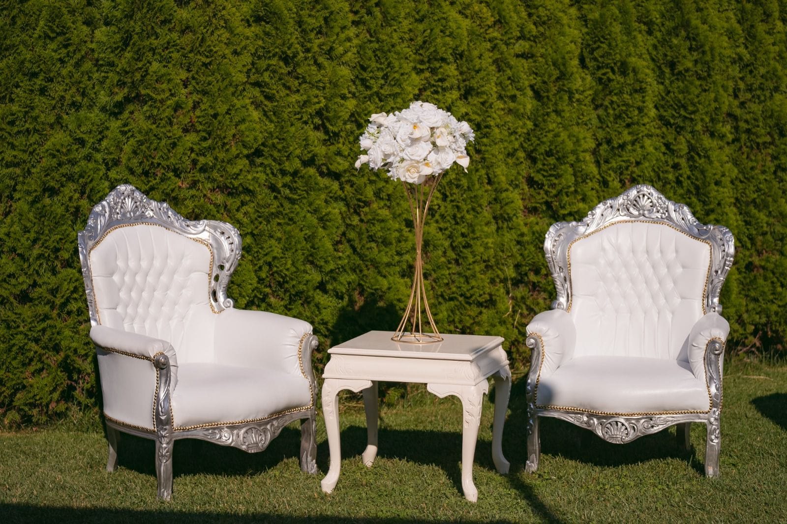 Deux chaises blanches richement décorées, ornées de motifs dorés, sont placées sur une petite table blanche sur laquelle se trouve un vase rempli de fleurs blanches. L’ensemble est mis en valeur par un arbuste taillé avec soin en arrière-plan.