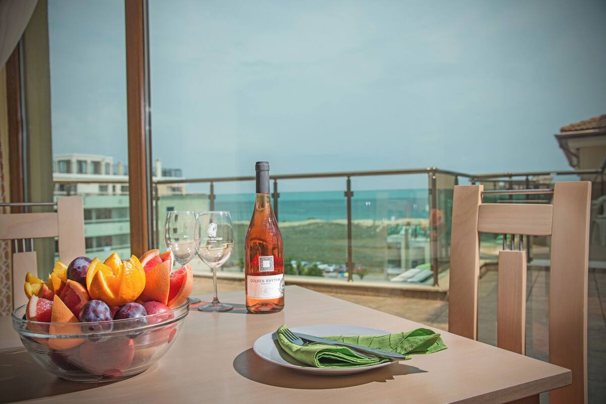 Una mesa con un frutero, una botella de vino y una copa en un balcón con vistas panorámicas.