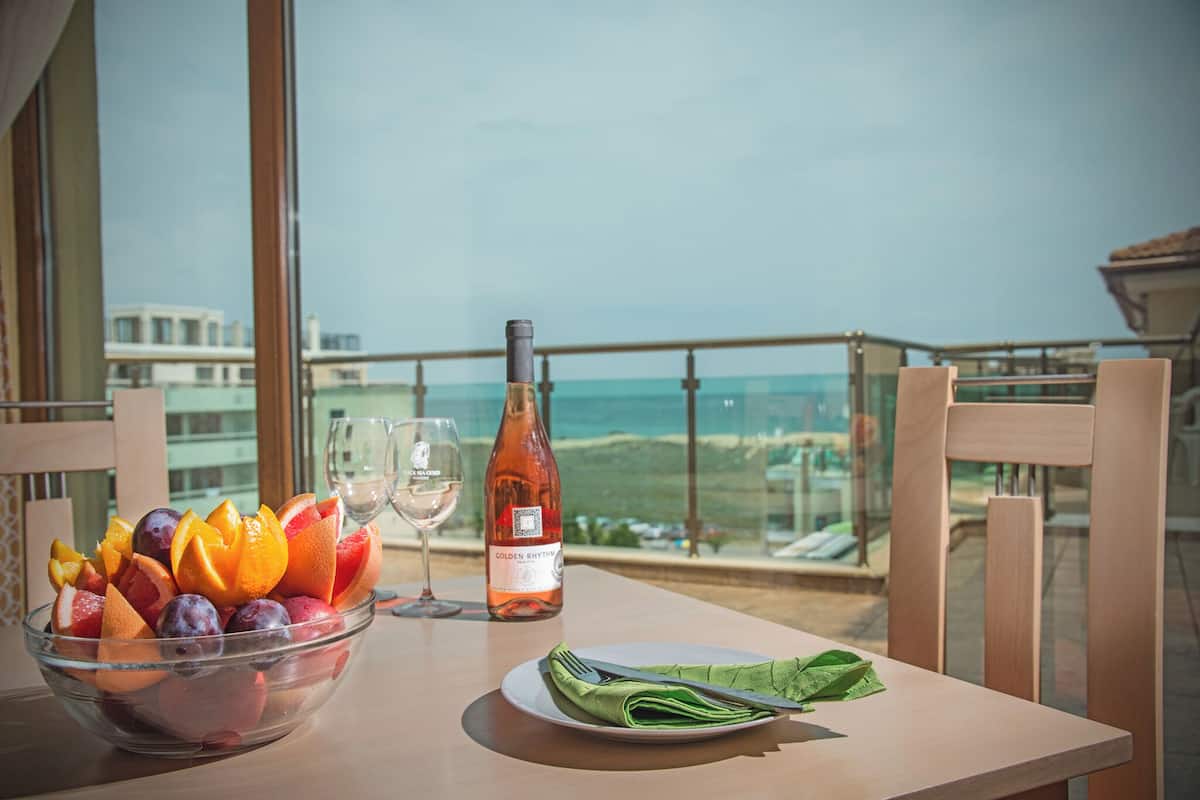 A table with a fruit bowl, a wine bottle, and a glass on a balcony overlooking a scenic view.