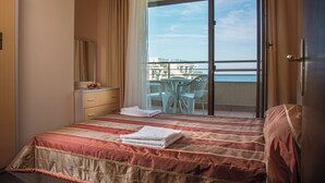 A hotel room with a bed, a desk with a chair, a balcony with a table and chairs, and a view of the sea.