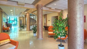 A spacious lobby with a central staircase, orange seating, and potted plants.