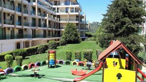 Un immeuble résidentiel à plusieurs étages doté de balcons, d’un terrain de jeux avec des toboggans et des structures ludiques, et d’une pelouse bien entretenue.