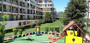 Un edificio residencial de varios pisos con balcones, un parque infantil con toboganes y juegos, y un césped bien cuidado.