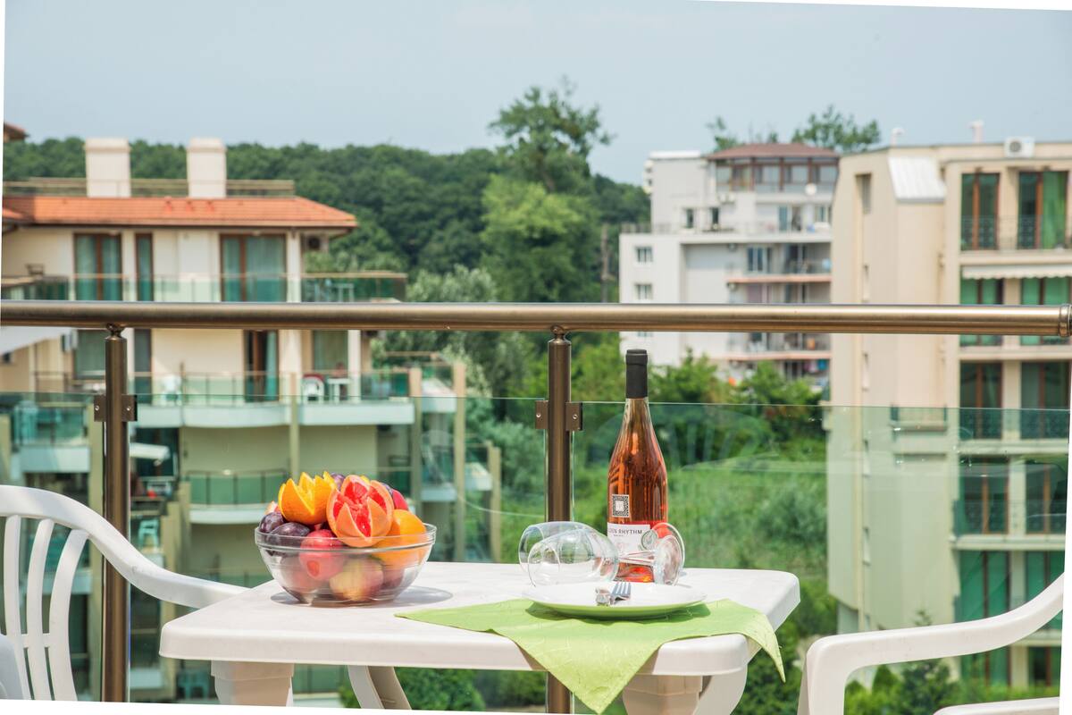 Un balcón con una mesa lista para dos personas, un frutero, una botella de vino y una copa.