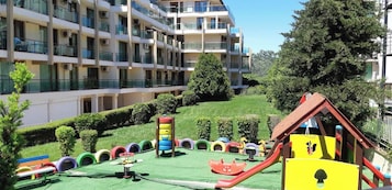 Un edificio residencial de varios pisos con balcones, un parque infantil con toboganes y juegos, y un césped bien cuidado.