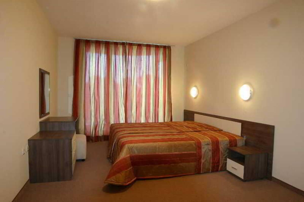 Una habitación de hotel con cama, mesitas de noche, espejo, cortinas y lámparas fijadas a la pared.