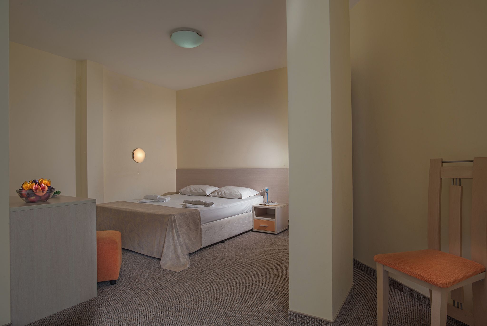 A hotel room with a bed, bedside table, chair, and a fruit bowl.