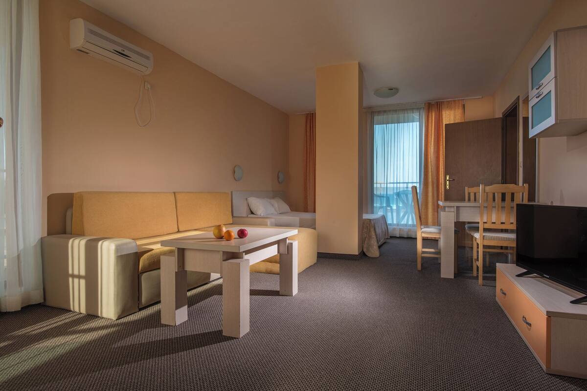 Habitación de hotel con sofá, mesa, televisión y balcón con cortinas.