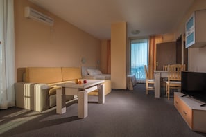 Habitación de hotel con sofá, mesa, televisión y balcón con cortinas.
