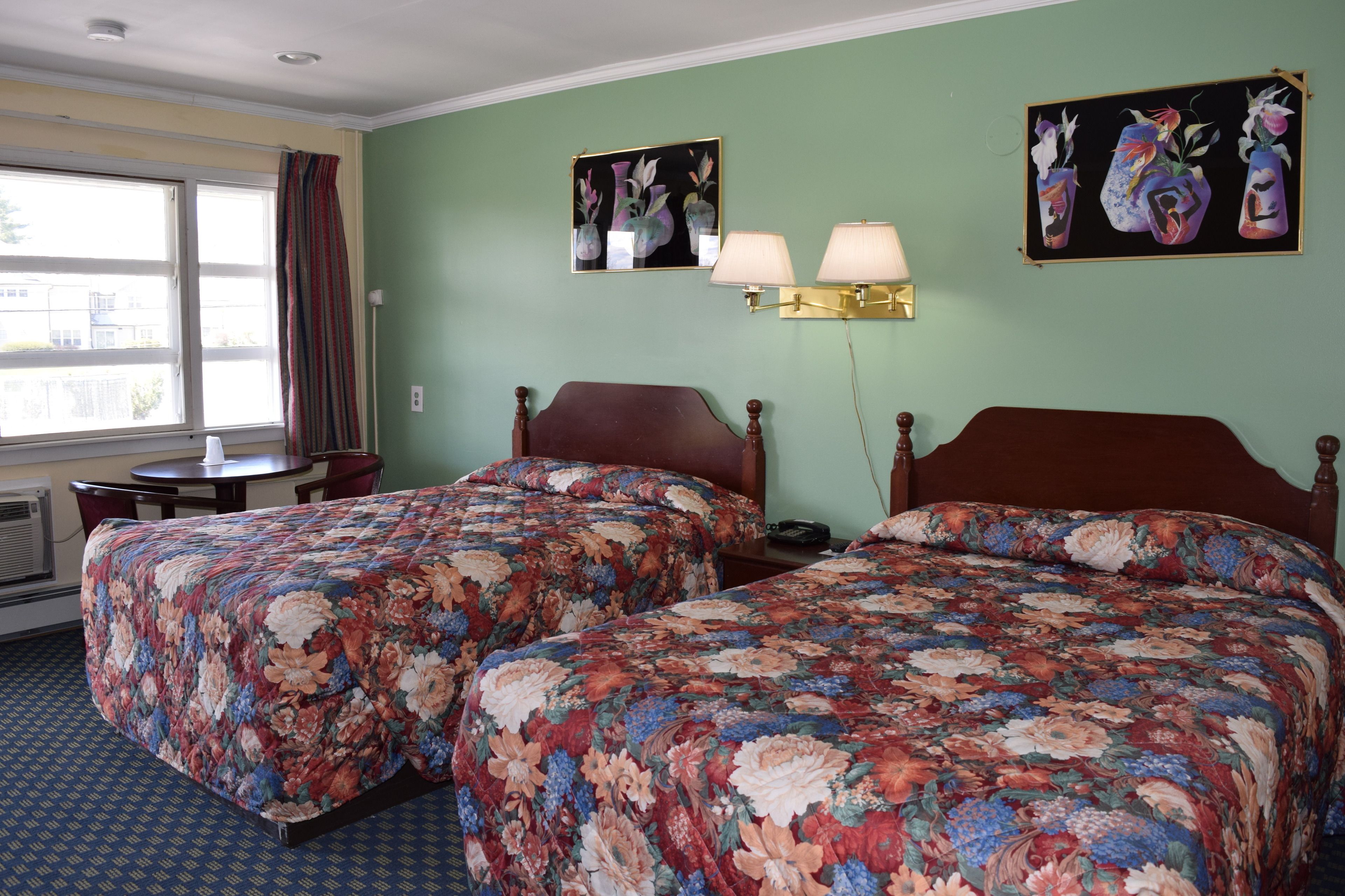 Una habitación de hotel con dos camas, cubrecamas estampados con motivos florales, una mesita de noche, una lámpara y obras de arte en las paredes.
