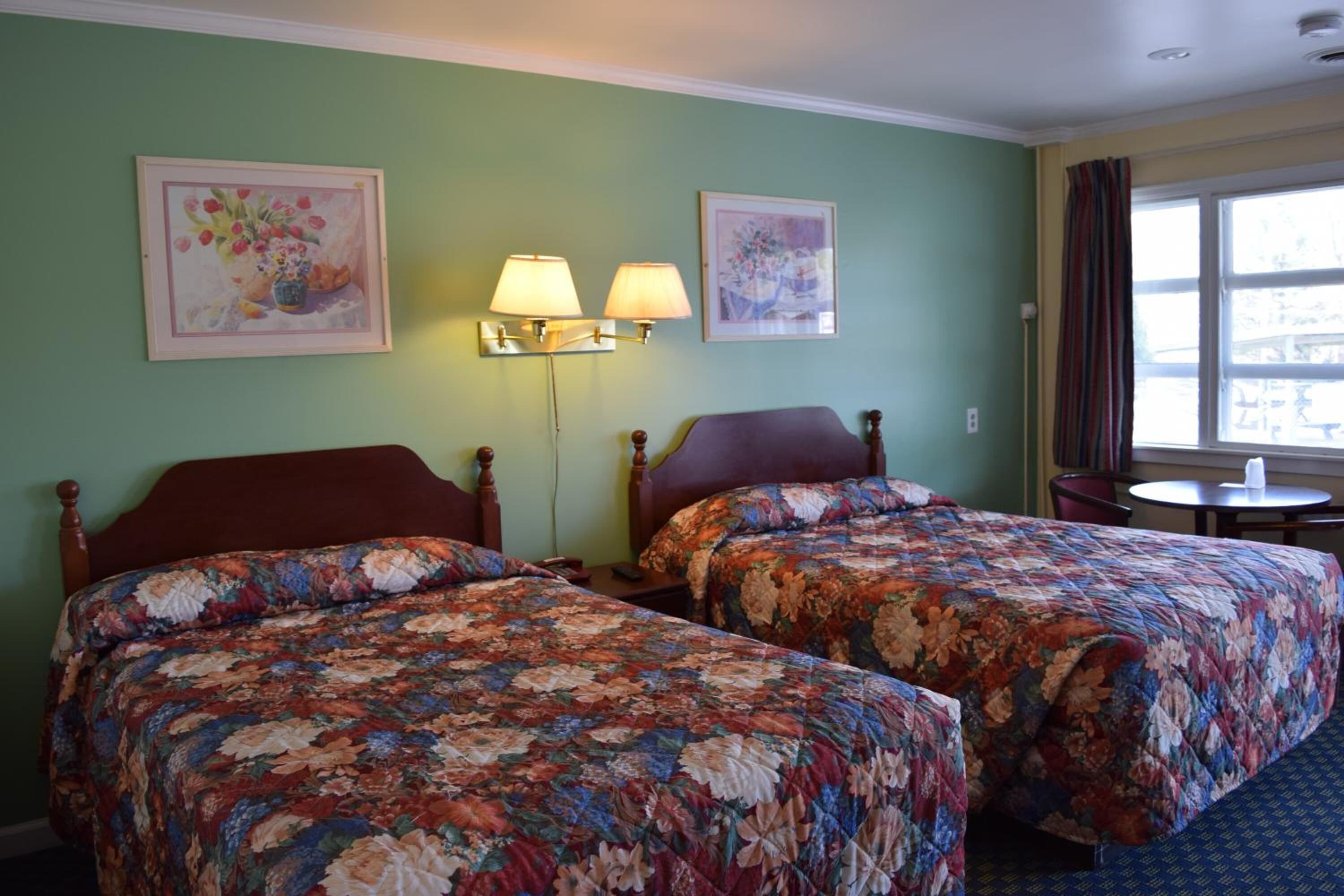 A hotel room with two beds, floral-patterned bedding, a small table, and two wall-mounted lamps.