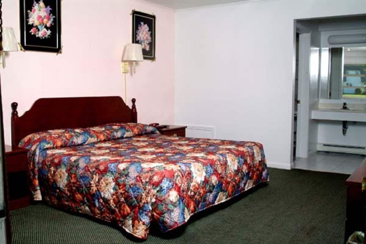 A bedroom with a floral bedspread, a wooden headboard, a nightstand, a lamp, and a bathroom visible through an open door.