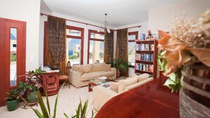 A living room with a sofa, a coffee table, a bookshelf, and potted plants. - Golden Sun (Karpathos)