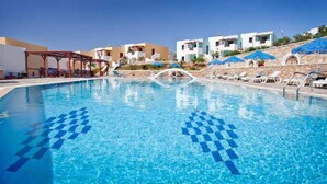 A swimming pool with clear blue water, lounge chairs, and a covered seating area. - Golden Sun (Karpathos)