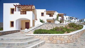 A white building with multiple windows and a stone staircase leading to a door. - Golden Sun (Karpathos)