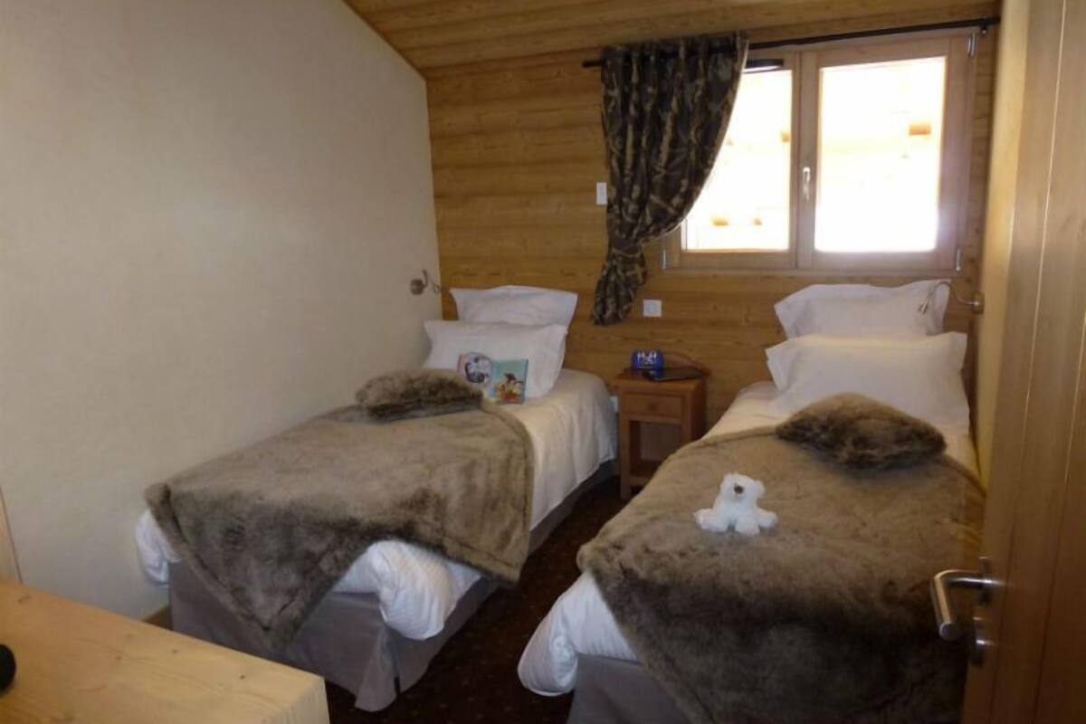 A room with two beds, wooden walls, a window with curtains, and a small bedside table.