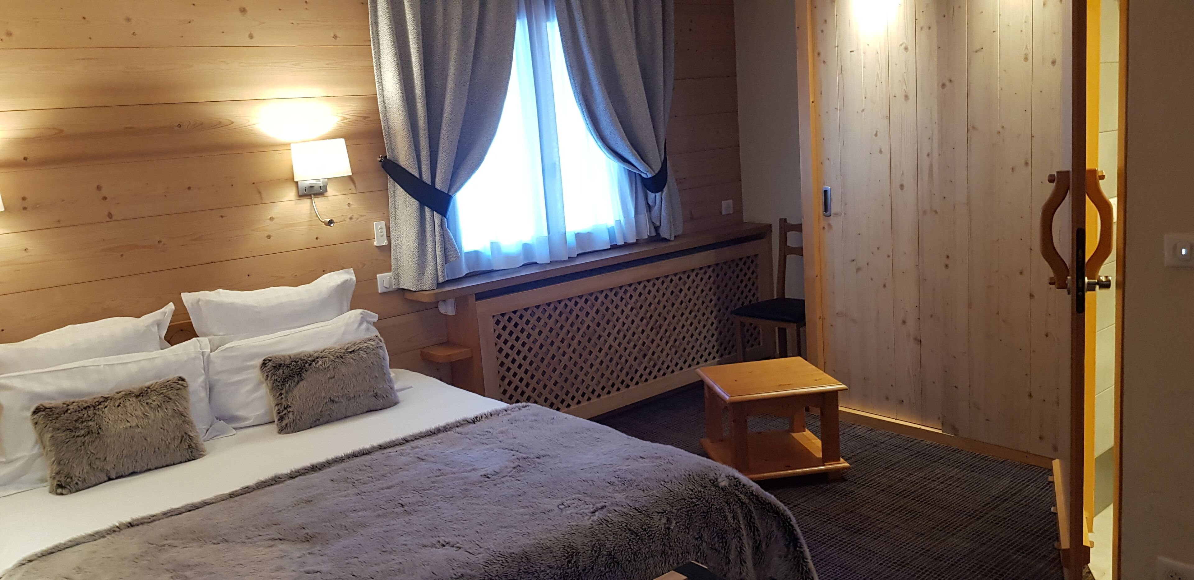 Una habitación con paneles de madera, una cama, un taburete pequeño y una ventana con cortinas.