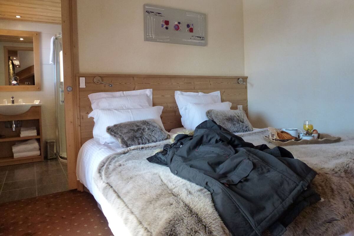 A hotel room with a wooden headboard, a large bed with white and grey pillows, a bathroom with a wooden vanity, and a wall-mounted artwork.