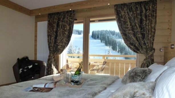 A bedroom with a large bed, a wooden headboard, a window with curtains, and a view of a snowy landscape. - Hôtel Restaurant Les Sapins Wellness & Gourmet (Manigod)