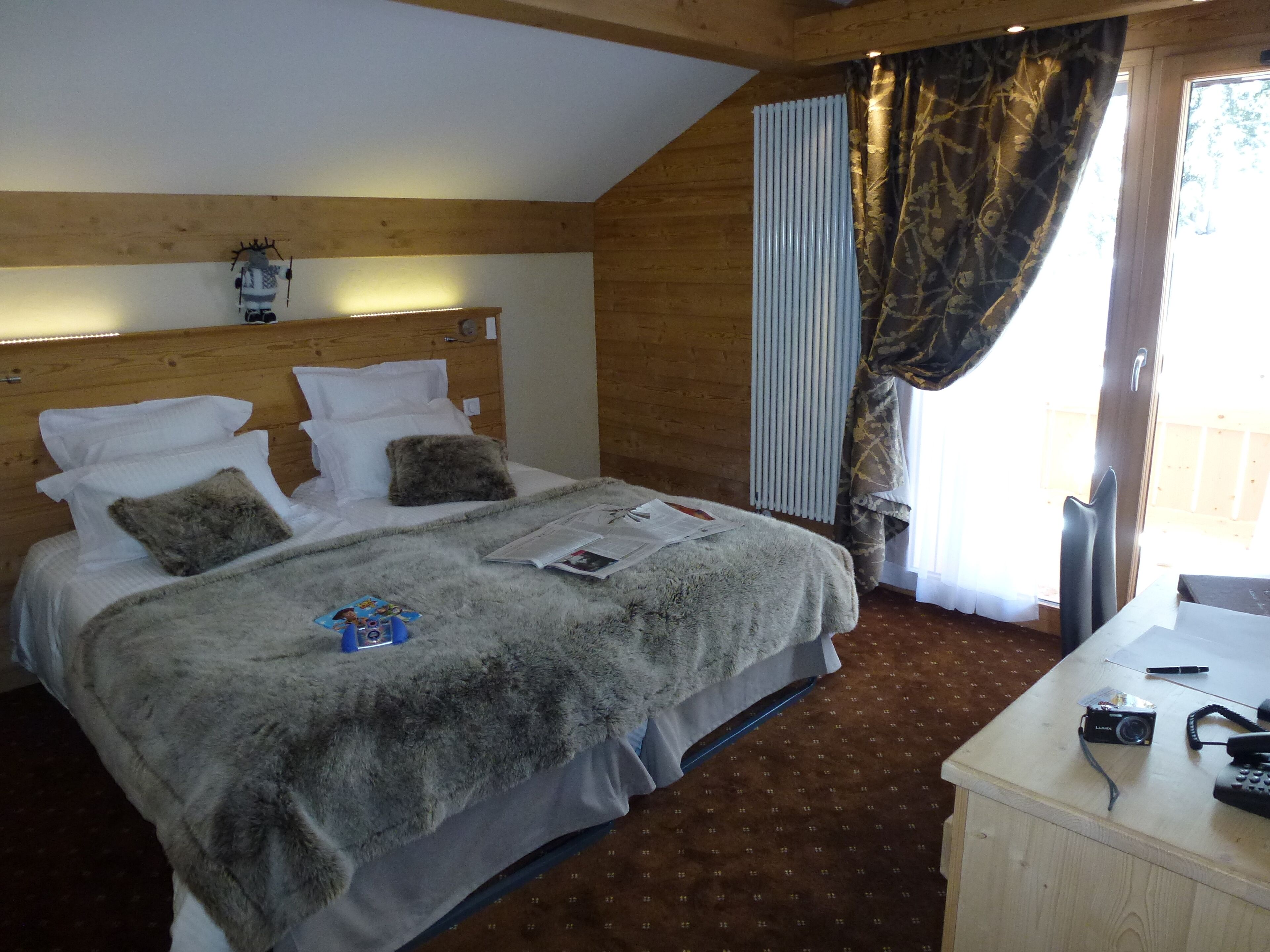 A hotel room with a wooden headboard, a bed with white linens and a grey furry blanket, a small table with a camera and phone, and a window with curtains.