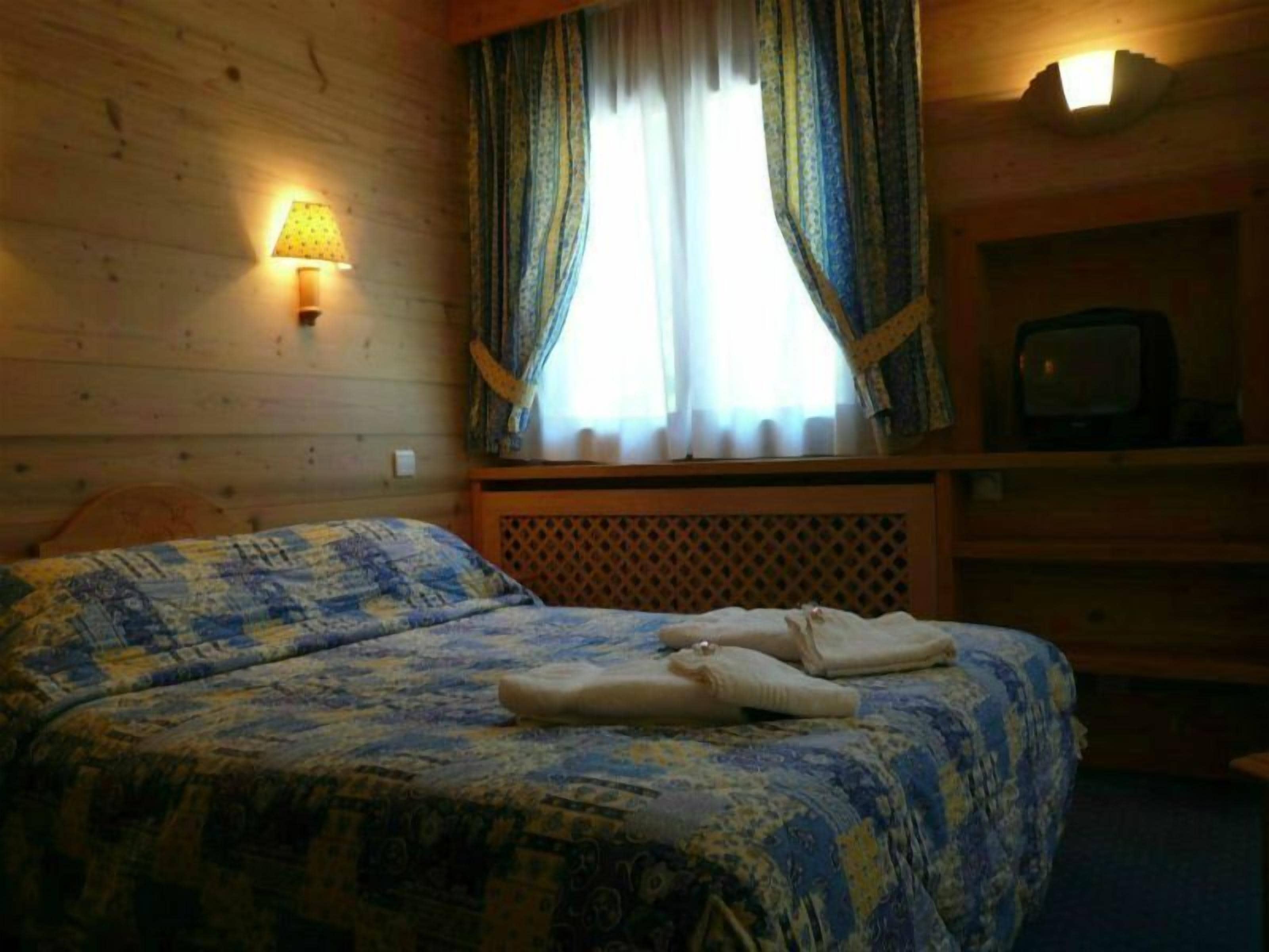 A room with two beds, a wooden headboard, a TV on a shelf, and a window with curtains.