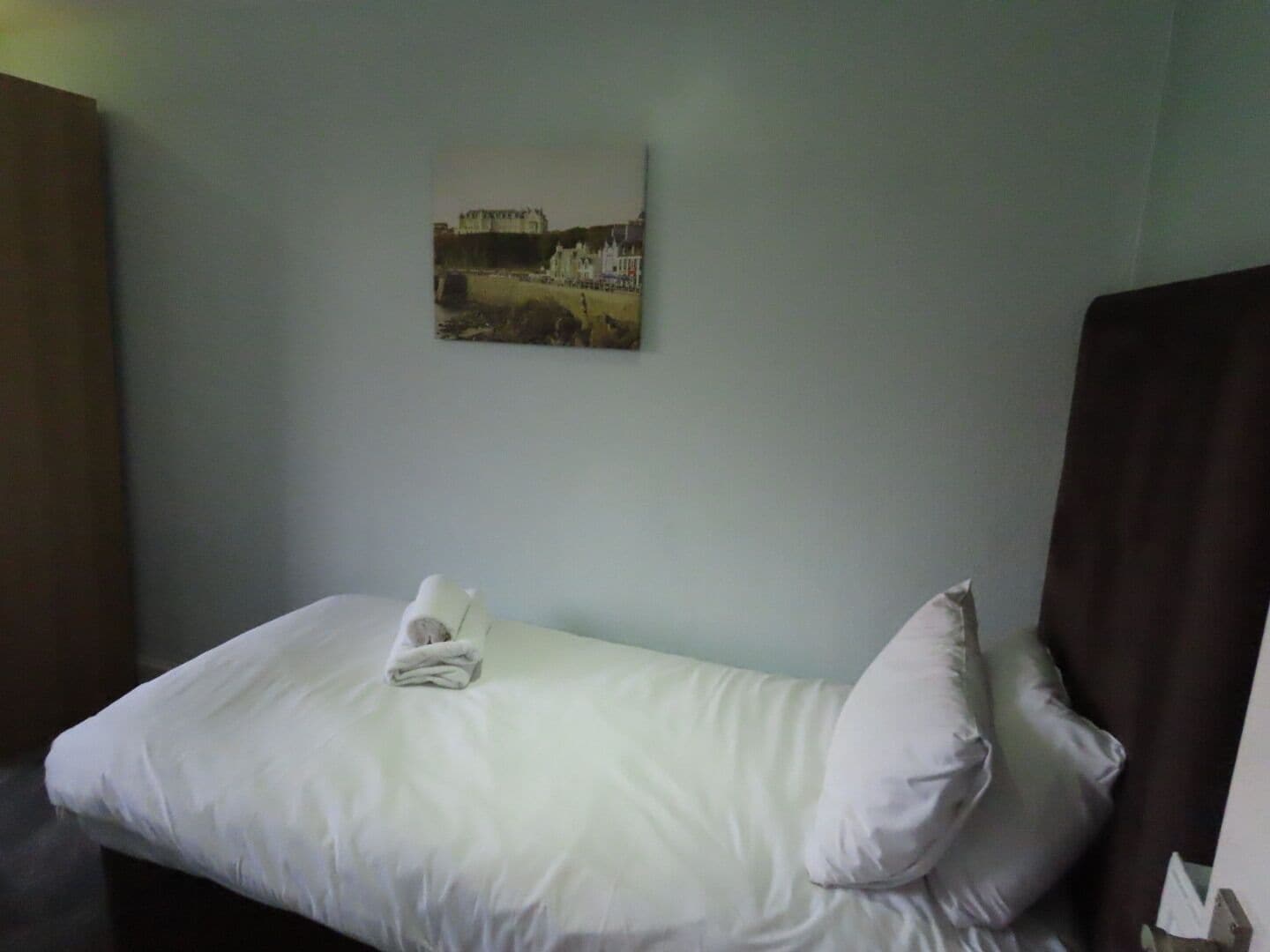 Een eenpersoonsbed met witte beddengoed, een opgevouwen handdoek en een kussen. Een lijst met een afbeelding van een kasteel hangt aan de muur.