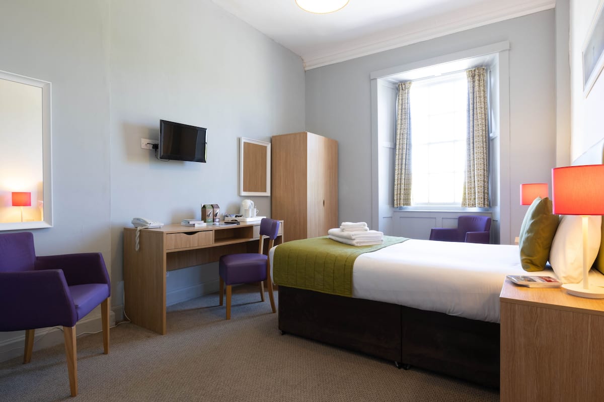 A hotel room with a bed, desk, chairs, a television, and a window with curtains.