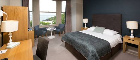 A hotel room with a large bed, a desk with a lamp, a chair, and a view of the sea.