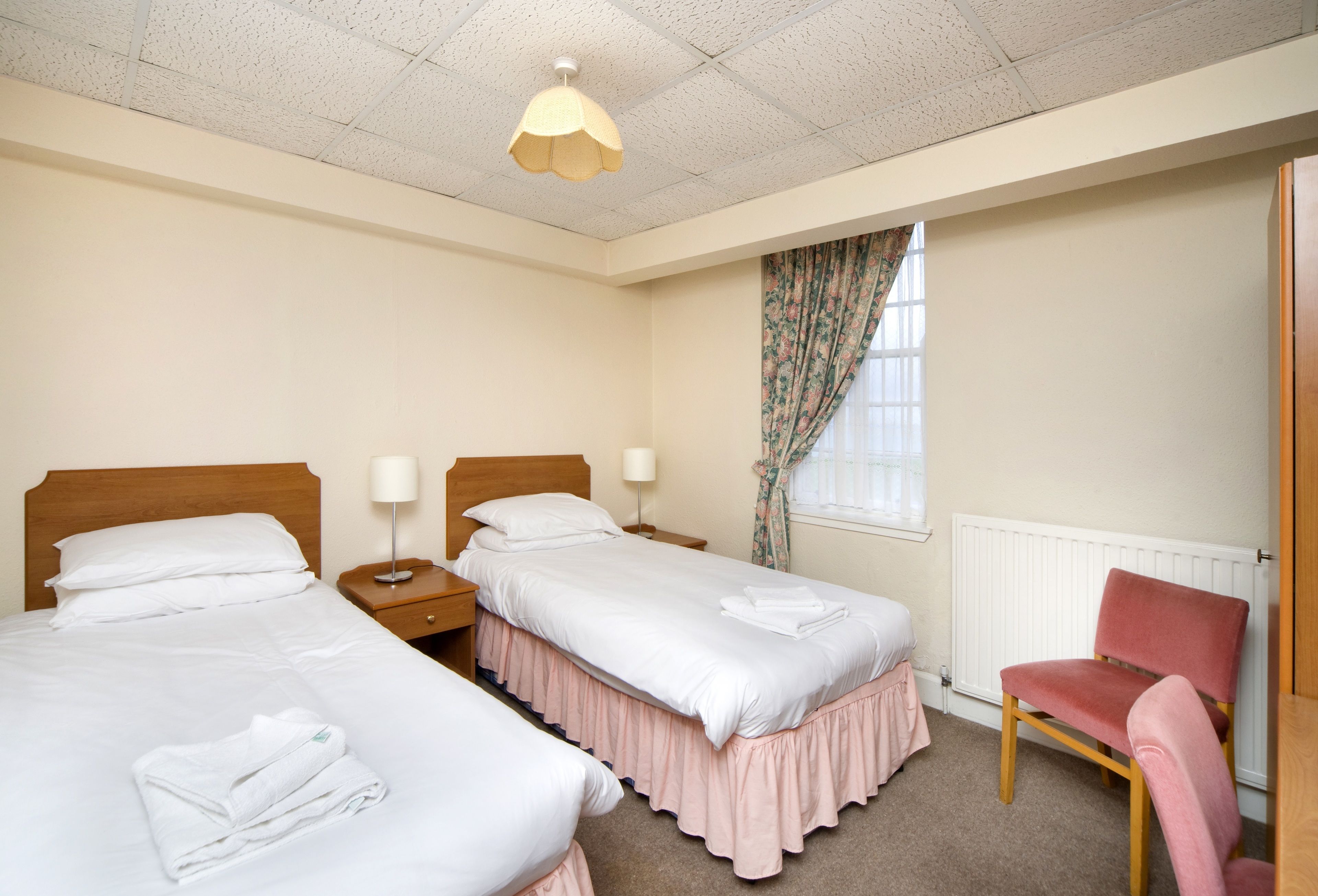 Een hotelkamer met twee bedden, een houten hoofdbord, een roze stoel, een radiator en een raam met bloemen gordijnen.