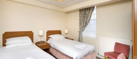 A hotel room with two beds, a wooden headboard, a pink chair, a radiator, and a window with floral curtains.