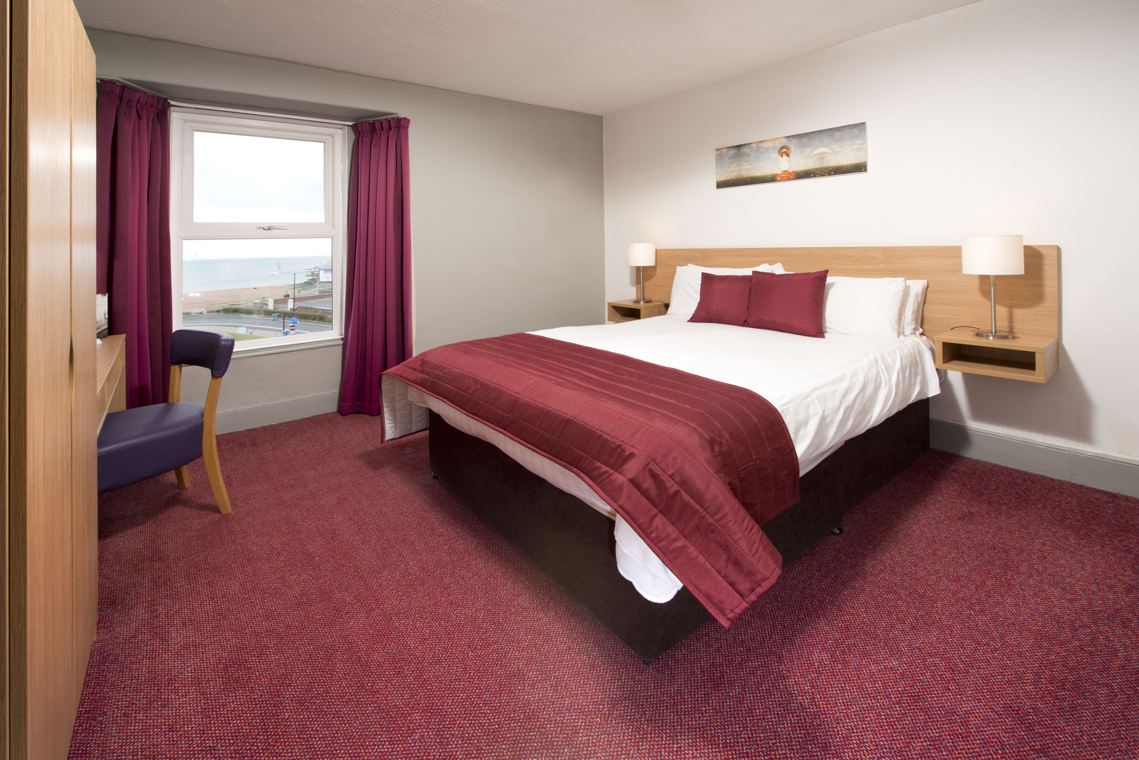 A hotel room with a large bed, a desk with a chair, a window with curtains, and a painting on the wall.