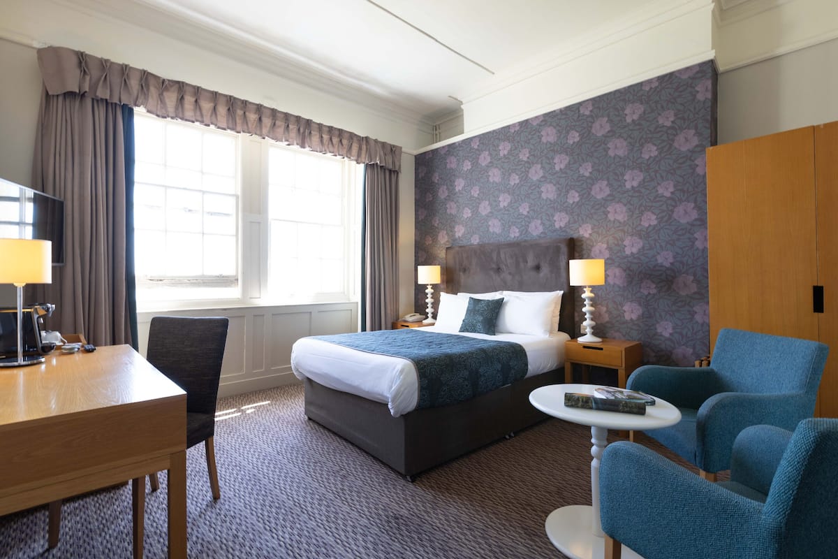 A hotel room with a large bed, two bedside tables with lamps, a desk, a chair, and a small round table.